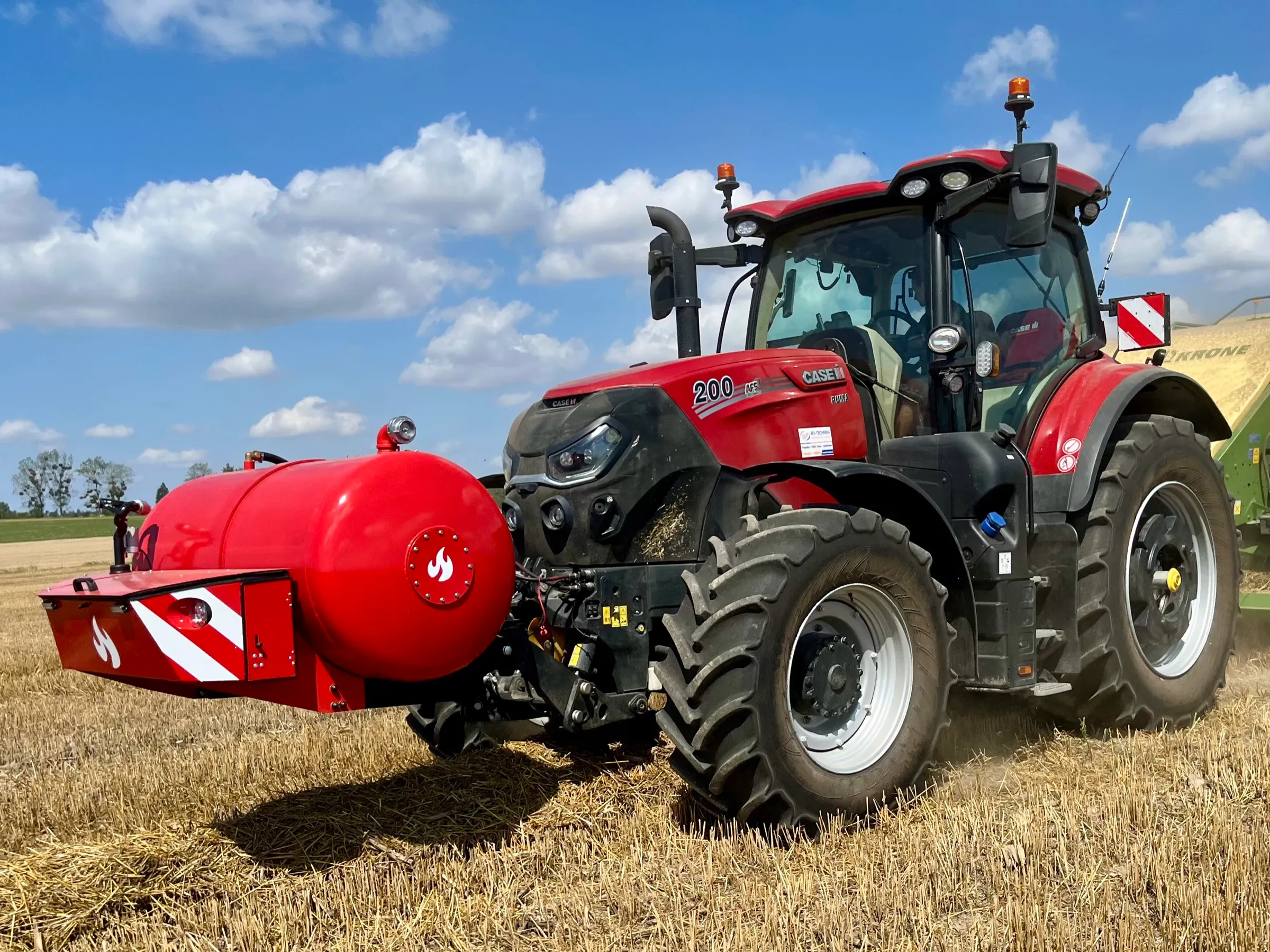 Traktor Case IH 200 s nádrží HYDROTANK na čelním závěsu na strništi pod modrou oblohou, vzadu lis