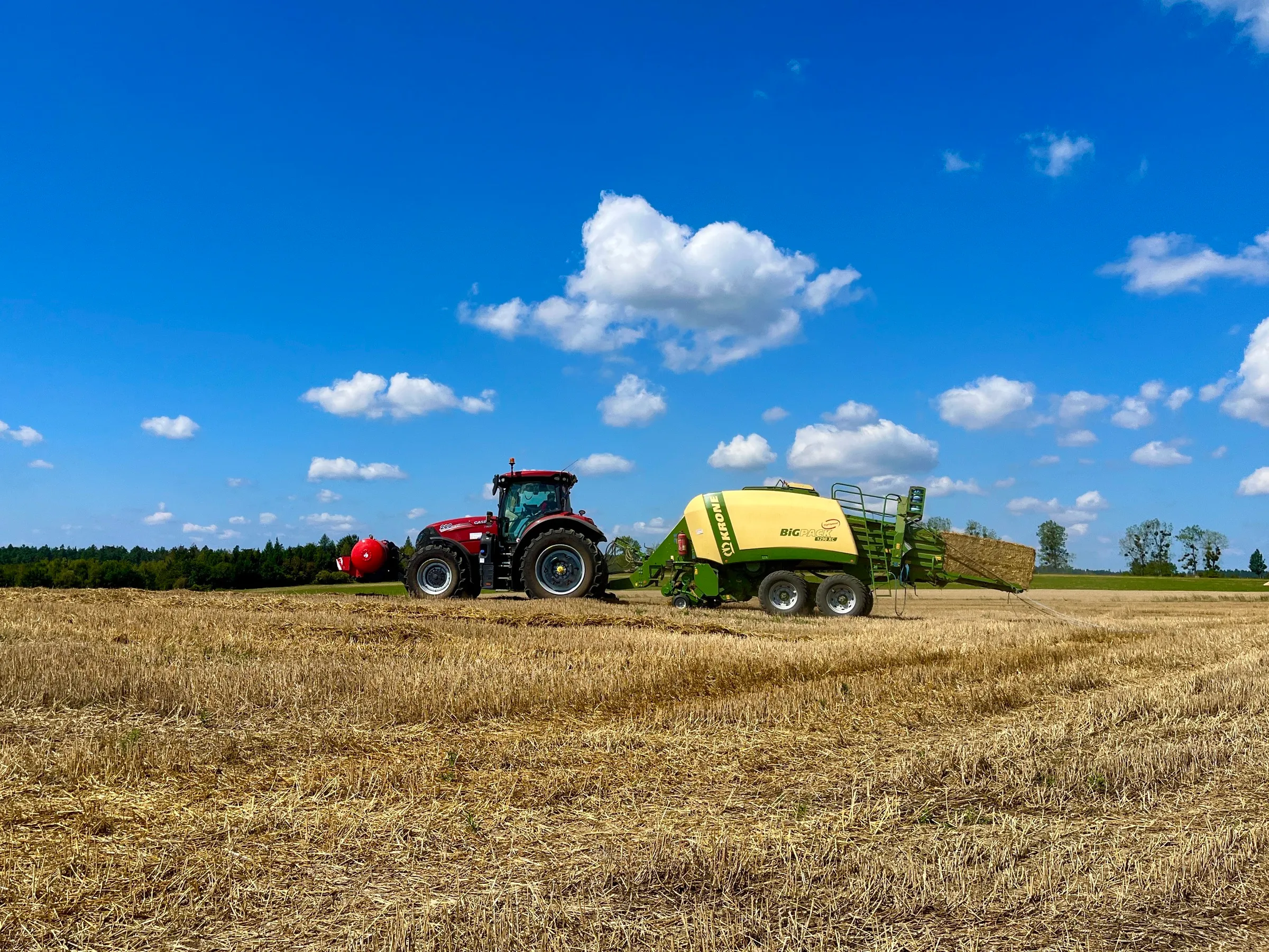 Traktor s HYDROTANKEM a velkoobjemovým lisem na rozlehlém strništi pod modrou oblohou